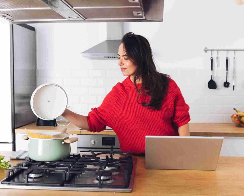 Woman cooking pasta on a built-in gas stove while using a laptop in a modern kitchen.
