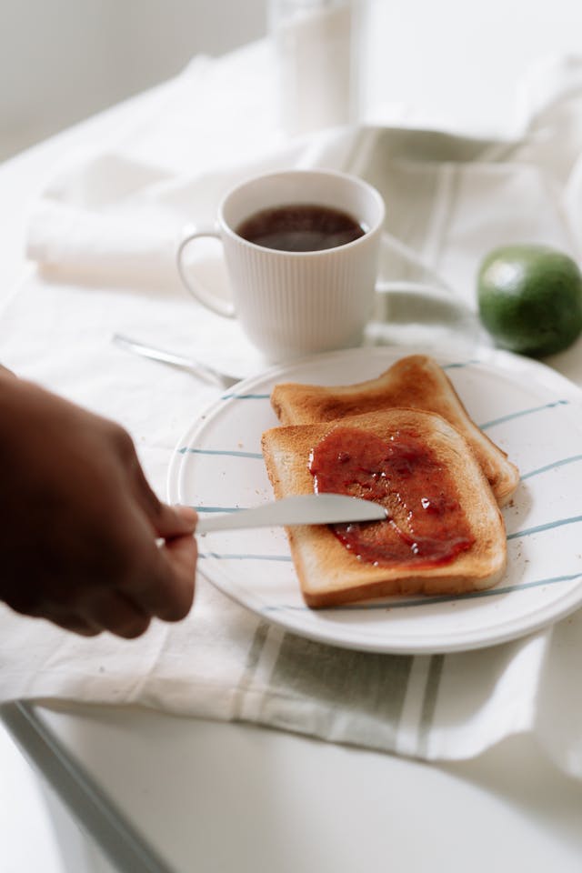 A hand spreading jam on toast beside a cup of coffee on a white plate.