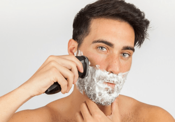 Man using an electric shaver with shaving foam on his face against a white background.
